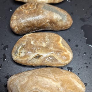 Puede incluir: Cuatro piedras lisas de forma ovalada en tonos marrones y beige. Las piedras tienen un aspecto pulido, con patrones y texturas visibles. La disposici&oacute;n es vertical, con las piedras ligeramente superpuestas.