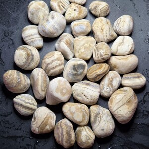 Pu&ograve; includere: Una collezione di pietre lisce e arrotondate nelle tonalit&agrave; del beige, crema e marrone. Le pietre presentano motivi e texture naturali, con alcuni che mostrano disegni a fasce o a strati. Le pietre sono disposte su una superficie scura.