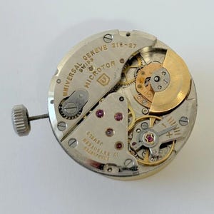 May include: Close-up of a watch movement with visible gears, jewels, and a winding stem. The words "UNIVERSAL GENEVE SWISS MICROROTOR" are inscribed on the metal plate. The movement features a gold-colored rotor and various other mechanical components.