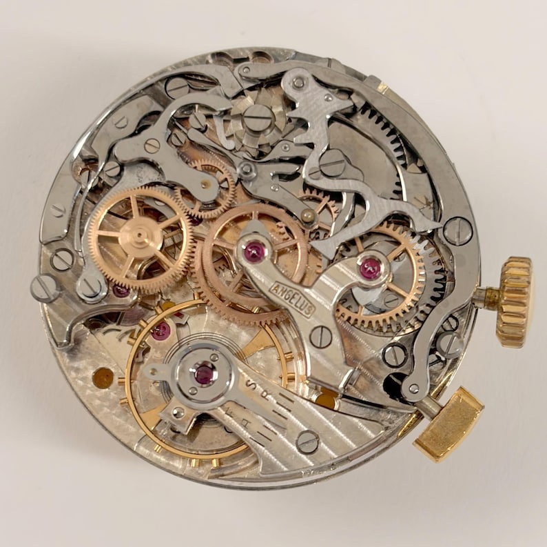 May include: Close-up of a watch movement with intricate gears, levers, and jewels. The mechanism features gold and silver components, with the word "ANGELUS" visible. A gold-colored crown and lever are on the right side.