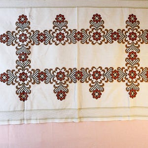 May include: White linen tablecloth with a detailed floral cross-stitch design in shades of red, brown, and black. The embroidered pattern forms a rectangular border with floral motifs at the corners and along the sides. The edges have a subtle striped trim.