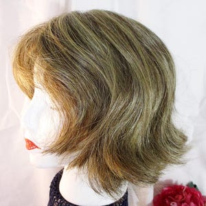 May include: A short, layered wig in shades of brown, blonde, and green, styled on a mannequin head. The wig features a side part and a slightly wavy texture. The mannequin has red lipstick.