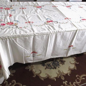 May include: A white rectangular tablecloth featuring intricate floral embroidery in shades of red, pink, and blue, accented with black scrollwork patterns. The fabric displays a smooth texture with visible folds and a decorative border design.