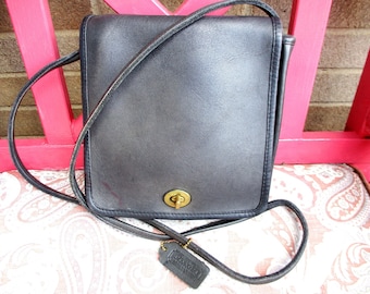 May include: A black leather crossbody bag with a gold-tone clasp and a long strap. The bag has a rectangular shape and a small leather tag attached. The bag is sitting on a patterned surface.