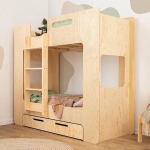 Op de afbeelding: Een lichtgekleurd houten stapelbed met een ladder, twee lades en een benedenbed. Het bovenste bed heeft een groene deken en een roze deken. Een kleine houten stoel en een plank staan ook in de kamer.