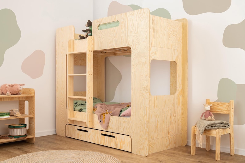 Op de afbeelding: Een lichtgekleurd houten stapelbed met een ladder, twee lades en een benedenbed. Het bovenste bed heeft een groene deken en een roze deken. Een kleine houten stoel en een plank staan ook in de kamer.
