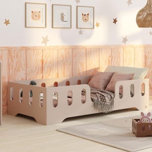 Puede incluir: Una cama de niño pequeña de color beige claro con una barandilla de seguridad y una manta blanca. La cama está en una habitación con un escritorio y una silla de madera, un triciclo de juguete de madera y una lámpara colgante de mimbre marrón.