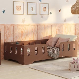Op de afbeelding: Een bruin houten peuterbed met een veiligheidshek, een wit en bruin dekbed en een paar kussens. Het bed staat in een kamer met een houten bureau en stoel, en een houten speelgoed-driewieler.