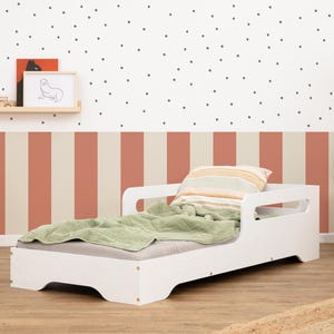 Op de afbeelding: Een wit peuterbed met een groene deken en gestreept kussen in een kinderkamer. De kamer heeft een gestreept wandontwerp in beige en koraal, met een gestippelde witte bovenmuur. Een houten plank bevat een ingelijste tekening.