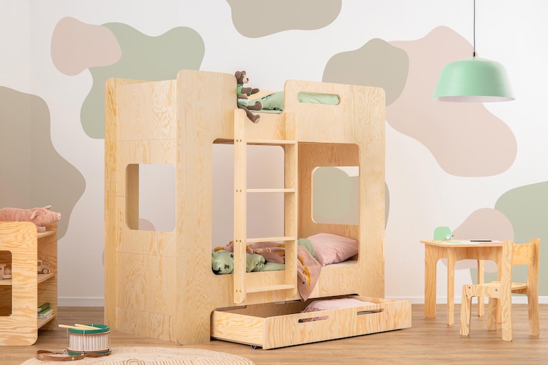 Op de afbeelding: Een lichtgekleurd houten stapelbed met een ingebouwde ladder, een uitschuifbare lade en een kleine tafel en stoel set. Het bed heeft een speels ontwerp met uitgesneden ramen en is versierd met knuffels en beddengoed.