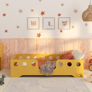 Könnte beinhalten: Ein gelbes Kinderbett mit runden Ausschnitten, Kissen und einer Decke. Der Raum zeigt Sternwandaufkleber, gerahmte Tierporträts sowie einen Schreibtisch und einen Stuhl aus Holz. Ein Spielzeugroller und ein Teppich sind ebenfalls zu sehen.