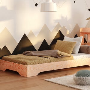 Op de afbeelding: Een licht houten platformbedframe met een geel en bruin gestreept deken en kussens. Het bed staat tegen een muur met een zwart-wit bergontwerp.