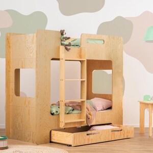 Op de afbeelding: Een lichtgekleurd houten stapelbed met een ingebouwde ladder, een uitschuifbare lade en twee slaapgedeeltes. Het bed heeft een eenvoudig, modern ontwerp en staat in een kamer met een neutraal kleurenpalet. Een kleine tafel en stoelset staan in de buurt.