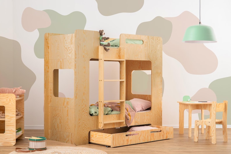 Op de afbeelding: Een lichtgekleurd houten stapelbed met een ingebouwde ladder, een uitschuifbare lade en twee slaapgedeeltes. Het bed heeft een eenvoudig, modern ontwerp en staat in een kamer met een neutraal kleurenpalet. Een kleine tafel en stoelset staan in de buurt.