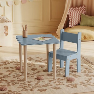 Op de afbeelding: Een lichtblauwe vierkante tafel met een geschulpte rand en vier lichthouten poten, gecombineerd met een bijpassende lichtblauwe houten stoel. Een kleine cilindrische pennenhouder met potloden en een vel papier staan op het tafelblad.