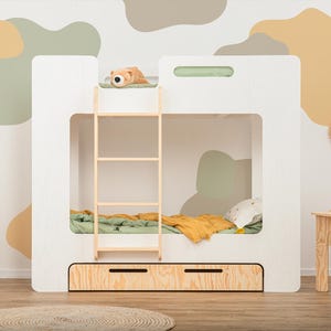 Op de afbeelding: Een wit stapelbed met een houten ladder en twee lades. Het bed heeft een groene en gele deken en een klein knuffeldier bovenop. De kamer heeft een neutraal kleurenpalet met abstracte muurkunst.