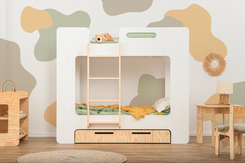 Op de afbeelding: Een wit stapelbed met een houten ladder en twee lades. Het bed heeft een groene en gele deken en een klein knuffeldier bovenop. De kamer heeft een neutraal kleurenpalet met abstracte muurkunst.
