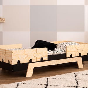 Könnte beinhalten: Ein Kinderbett aus Holz mit schwarz lackierten Seiten und einem integrierten Sicherheitsgitter. Das Bett hat eine Matratze mit weiß-schwarzem Muster und ein Kissen mit weiß-schwarzem Muster.