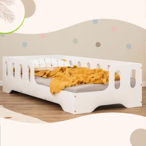 Puede incluir: Una cama para niños pequeños de madera blanca con un perfil bajo y esquinas redondeadas. La cama está hecha con un colchón gris, una almohada blanca con puntos negros y una manta de color amarillo mostaza. La cama está colocada contra una pared neutra con lunares de colores.