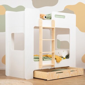 Op de afbeelding: Een wit stapelbed met een houten ladder en een uitschuifbare lade. Het bed heeft een minimalistisch ontwerp met rechthoekige uitsparingen. De kamer heeft een houten bijzettafel, een kleine stoel en een decoratieve wandophanging. De muur is beschilderd met abstracte vormen.