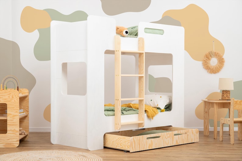 Op de afbeelding: Een wit stapelbed met een houten ladder en een uitschuifbare lade. Het bed heeft een minimalistisch ontwerp met rechthoekige uitsparingen. De kamer heeft een houten bijzettafel, een kleine stoel en een decoratieve wandophanging. De muur is beschilderd met abstracte vormen.