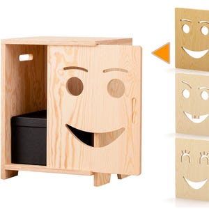 Op de afbeelding: Een lichtgekleurde houten kast met een lachend gezichtsontwerp op de deur. De kast heeft een rechthoekige vorm met een opbergvak en een zwarte doos binnenin. Extra deuren met verschillende gezichtsuitdrukkingen worden getoond.