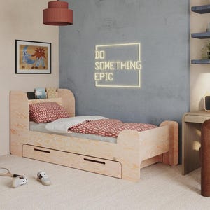 Può includere: Un letto in legno chiaro con due cassetti, una testiera con ripiano e un cuscino e copriletto a fantasia rossa. La stanza comprende una scrivania, una sedia e un tavolino blu. Il muro ha un'insegna al neon che dice "DO SOMETHING EPIC".