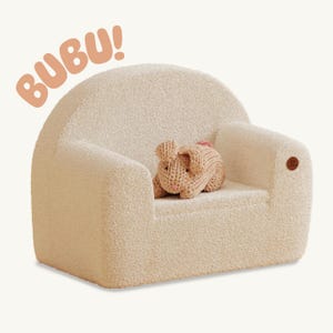Può includere: Poltroncina per bambini color crema con schienale e braccioli arrotondati. Un giocattolo a forma di coniglio beige lavorato a maglia con accenti rosa è seduto sul sedile. La parola "BUBU!" è scritta in arancione sopra la sedia.