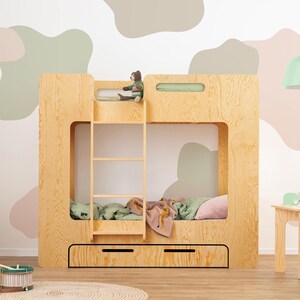 Op de afbeelding: Een houten stapelbed met een ladder, lades en een uitgesneden ontwerp. Het bed staat in een kamer met een bijpassend klein tafeltje en stoel. Een groene en roze deken en kussens liggen op het onderste stapelbed.