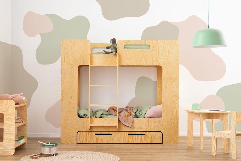 Op de afbeelding: Een houten stapelbed met een ladder, lades en een uitgesneden ontwerp. Het bed staat in een kamer met een bijpassend klein tafeltje en stoel. Een groene en roze deken en kussens liggen op het onderste stapelbed.