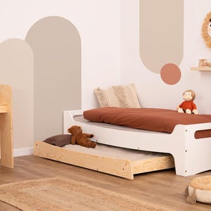 Pode incluir: Estrutura de cama de madeira branca com um colchão castanho e uma gaveta retrátil. A cama é decorada com um cobertor de lã castanho, um animal de peluche e uma almofada com um padrão geométrico. A parede atrás da cama é pintada com um esquema de cores castanho claro e bege com formas abstratas.