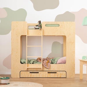Op de afbeelding: Een lichtgekleurd houten stapelbed met een ladder, twee lades en een klein berenspeeltje bovenop. De kamer heeft een lichtgroene hanglamp en een kleine houten tafel en stoel. De muur is versierd met abstracte vormen in neutrale kleuren.
