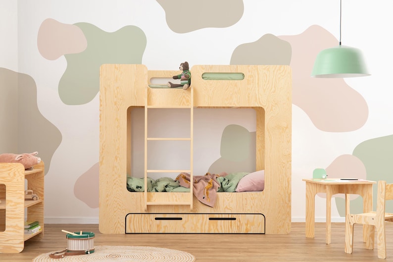 Op de afbeelding: Een lichtgekleurd houten stapelbed met een ladder, twee lades en een klein berenspeeltje bovenop. De kamer heeft een lichtgroene hanglamp en een kleine houten tafel en stoel. De muur is versierd met abstracte vormen in neutrale kleuren.