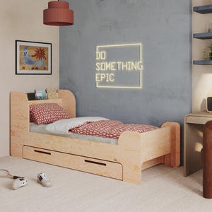 Puede incluir: Una cama infantil de madera clara con cabecero, cajones de almacenamiento y una colcha con estampado rojo. La habitación cuenta con un escritorio, una silla y un letrero de pared que dice "DO SOMETHING EPIC". También se ve un armario azul y obras de arte.