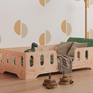 Può includere: Un lettino in legno chiaro per bambini piccoli con bordi arrotondati e dettagli ritagliati. Il letto è arredato con cuscini, una coperta e un giocattolo a forma di dinosauro verde. Sono visibili anche un'auto giocattolo in legno e giocattoli impilabili.