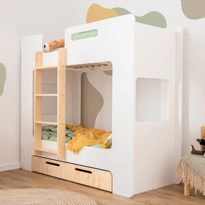 Op de afbeelding: Een wit stapelbed met een houten ladder en een uitschuifbare lade. Het bed heeft een groene en gele deken en een knuffel bovenop. De kamer heeft een houten stoel, een kleine houten plank en een vloerkleed.