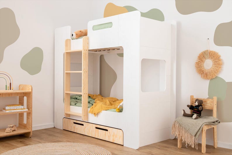 Op de afbeelding: Een wit stapelbed met een houten ladder en een uitschuifbare lade. Het bed heeft een groene en gele deken en een knuffel bovenop. De kamer heeft een houten stoel, een kleine houten plank en een vloerkleed.