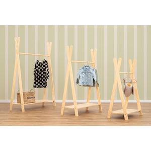 Op de afbeelding: Drie houten kledingrekken in tipi-stijl met kleding en een canvas tas. De rekken zijn gemaakt van lichtgekleurd hout en hebben een plank aan de onderkant. Een zwarte jurk met witte bloemen, een spijkerjasje en een knuffel worden getoond.