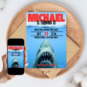 Op de afbeelding: Een blauwe en rode verjaardagsuitnodiging met een haaienvin en tanden die uit het water komen. De tekst luidt "Michael wordt 10. Doe mee met een Jaws-some zwembadfeest. Juli 24, 12 uur. Michaels huis | 123 Ocean Street. RSVP Jillian 123-456-7890. 5" x 7"