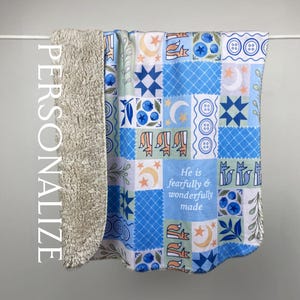 Può includere: Una coperta patchwork blu e bianca con una morbida fodera in sherpa beige. La coperta presenta un motivo ripetuto di stelle, lune, volpi e mirtilli. Il testo "He is fearfully & wonderfully made" è visibile.