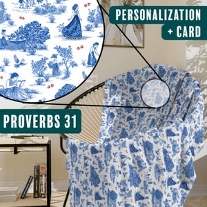 May include: A white throw blanket with a blue and white floral pattern. The blanket is folded and draped over a chair. The blanket has the text "Proverbs 31" on it. The image also includes a sign that says "Personalization + Card".