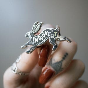 May include: A silver-colored enamel pin in the shape of a running rabbit. The word "Introvert" is written across the rabbit's body in white lettering. The pin has black and light yellow accents.