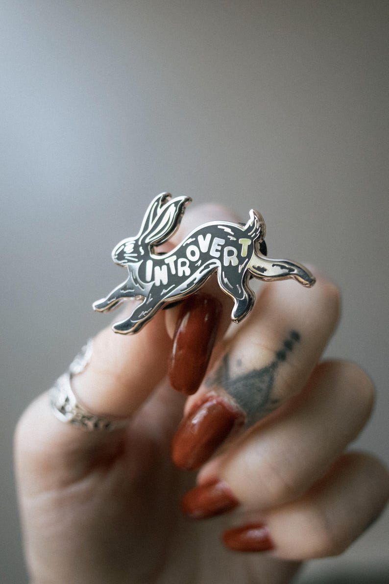 May include: A silver-colored enamel pin in the shape of a running rabbit. The word "Introvert" is written across the rabbit's body in white lettering. The pin has black and light yellow accents.