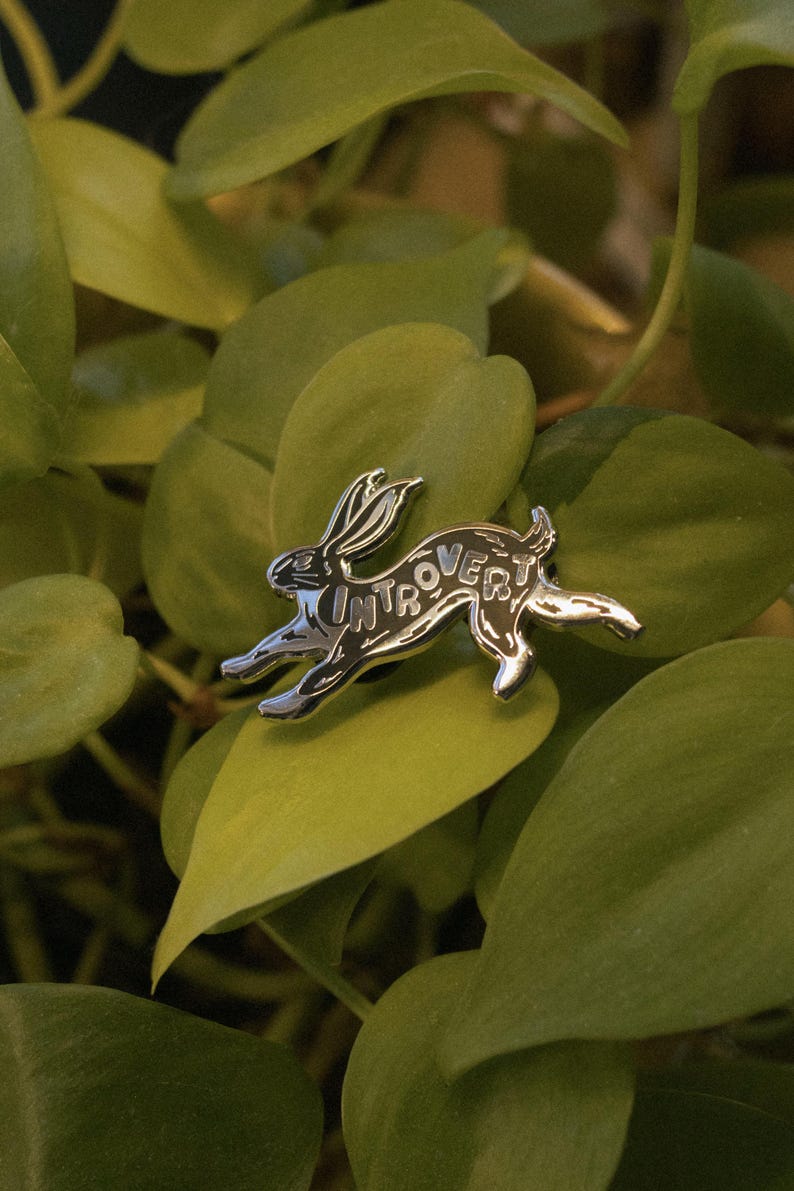 May include: A silver-colored pin in the shape of a running rabbit. The word "INTROVERT" is inscribed on the rabbit's body. The pin is set against a backdrop of green leaves, creating a nature-inspired aesthetic.
