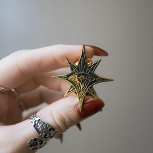 May include: A gold-colored star-shaped pin with a black enamel design. The pin features an eye and abstract shapes within the star's points. The pin is held between fingers, showcasing its intricate details and craftsmanship.
