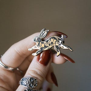 May include: A black and gold enamel pin in the shape of a running rabbit. The word "Introvert" is written across the rabbit's body. The pin is held by a person with long, red nails and silver rings.