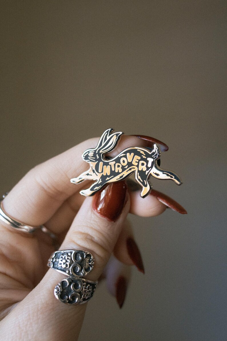 May include: A black and gold enamel pin in the shape of a running rabbit. The word "Introvert" is written across the rabbit's body. The pin is held by a person with long, red nails and silver rings.