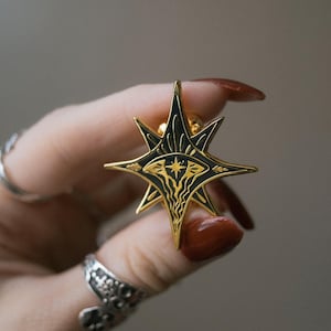 May include: A gold-colored star-shaped pin with a black and gold design. The pin features a central eye design and intricate patterns. The pin is held in a hand with long, polished nails and silver rings.