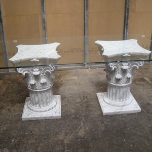 Op de afbeelding: Een tafel met glazen blad en twee witte, zuilvormige sokkels. Elke sokkel heeft een vierkante basis, een gegroefde kolom en een decoratief kapiteel. Een uniek meubelstuk.