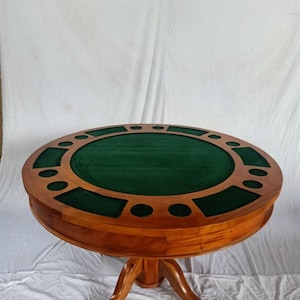 Puede incluir: Una mesa de póquer redonda de madera con una superficie de juego de fieltro verde. La mesa tiene un acabado de madera oscura y cuenta con posiciones para jugadores con portavasos. La mesa está soportada por una base.
